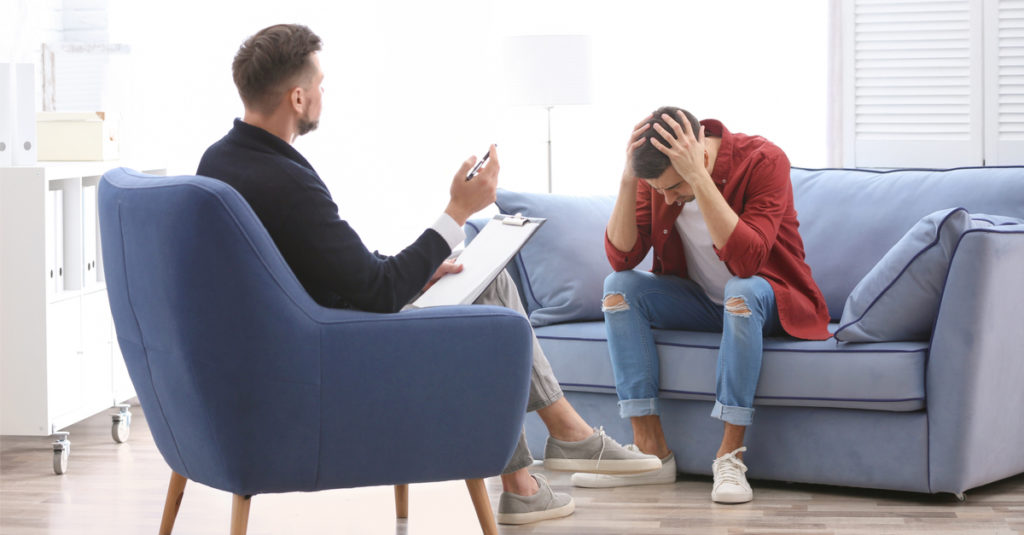 pain and suffering male on sofa stressed with hands on head while speaking with his male therapist